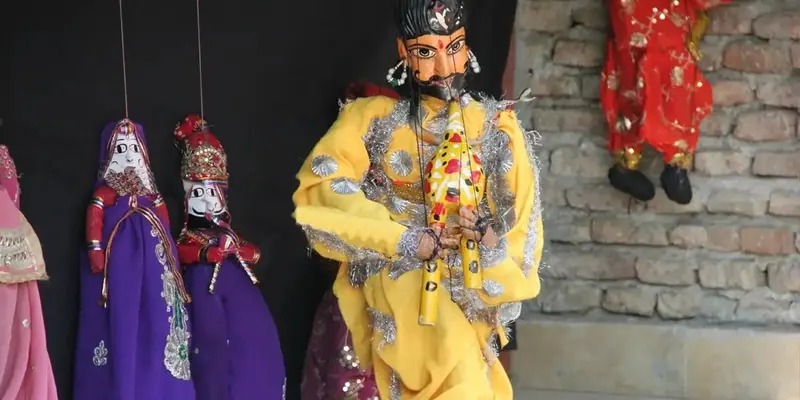 Yellow-robed string puppet with painted face beside smaller purple and red puppets on a brick wall