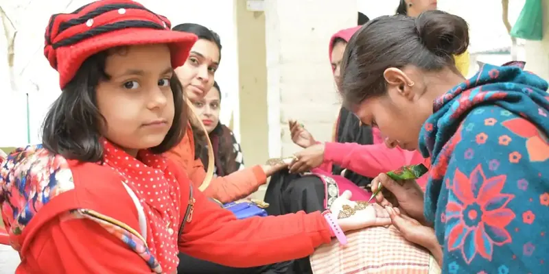 Girl in a red cap watches a mehndi artist apply henna to her outstretched hand