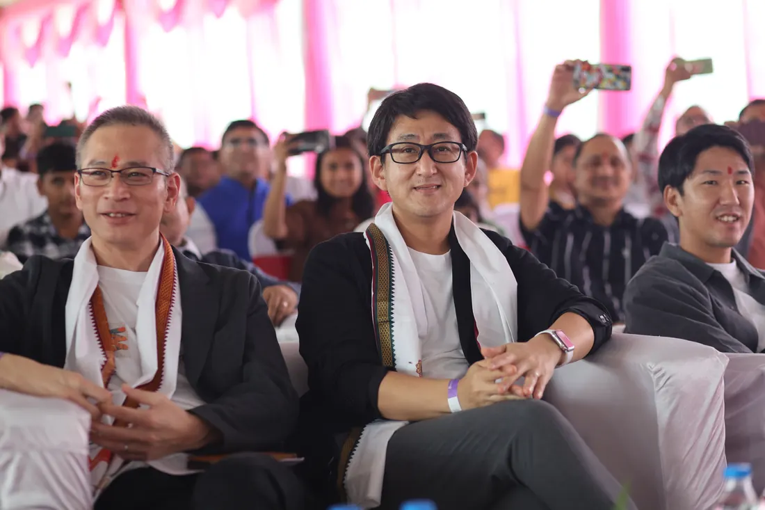 Seated corporate guests wearing garlands and tilak, watching a programme under a pink-draped pandal