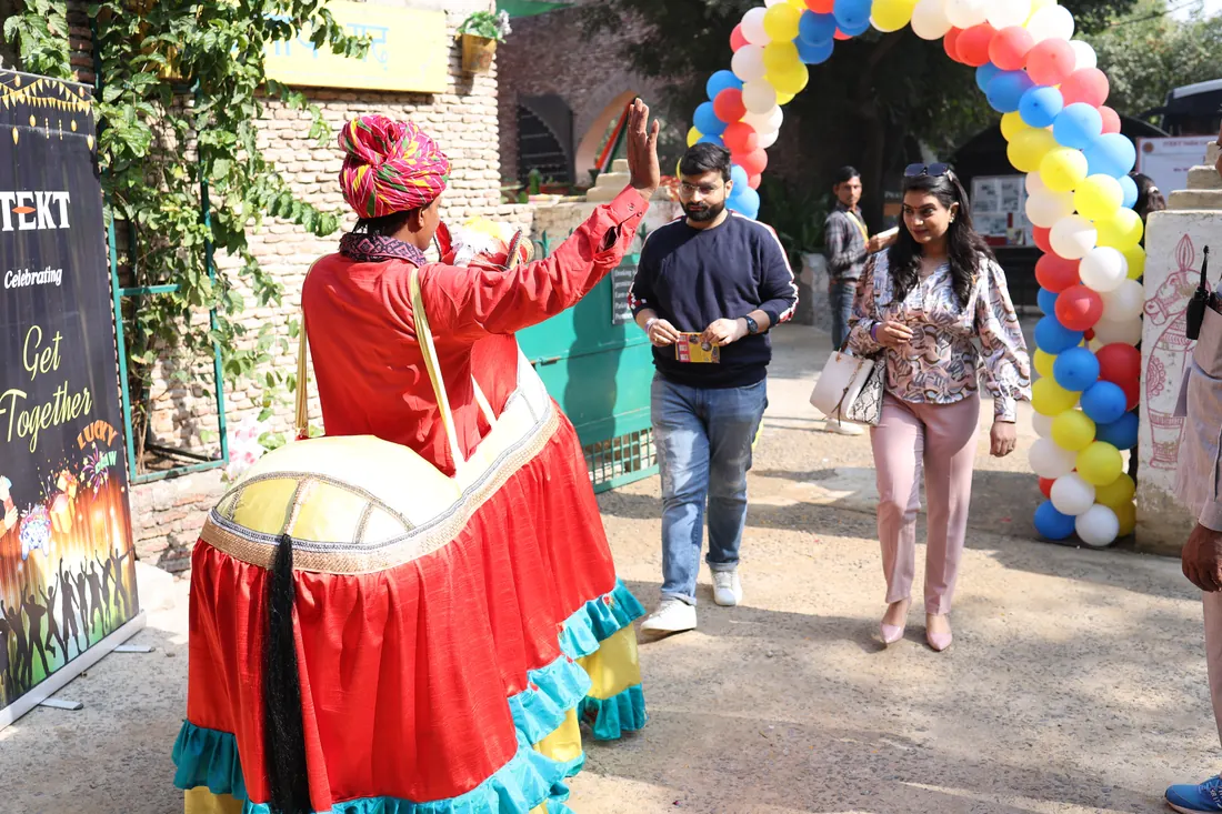 Folk artist in a red turban and bright skirt welcoming a guest with a raised hand at the balloon-decorated entrance
