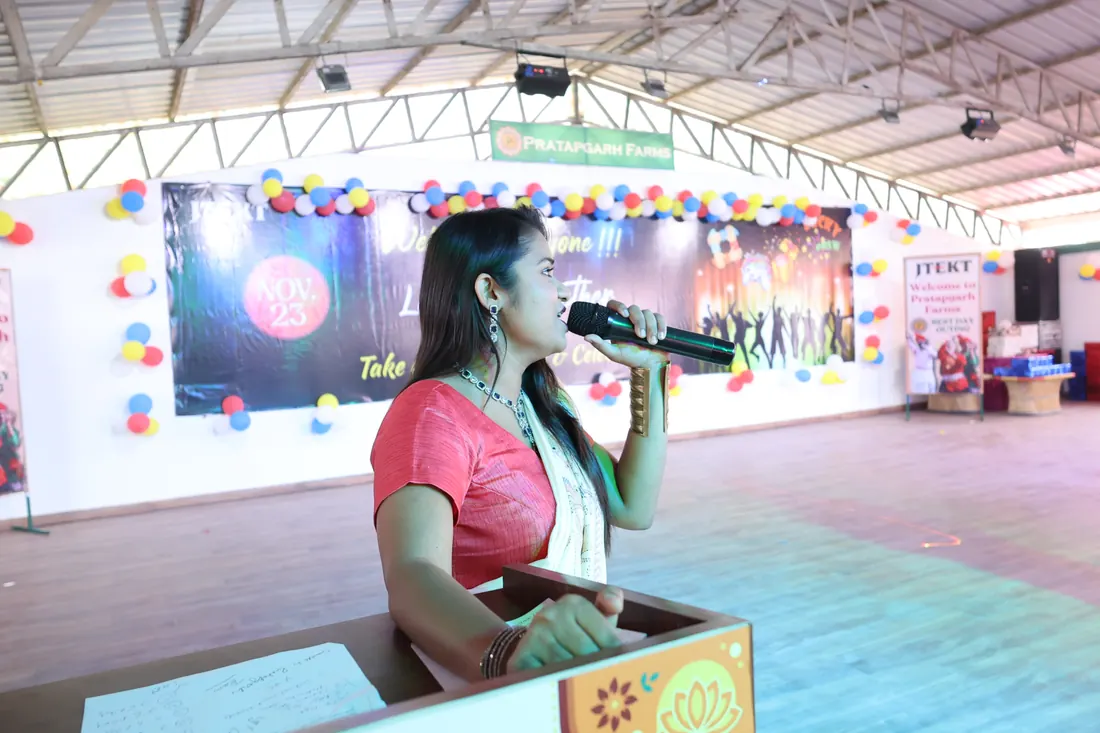 Woman speaking into a microphone at a wooden podium on stage, with a Pratapgarh Farms banner and a get-together backdrop behind her