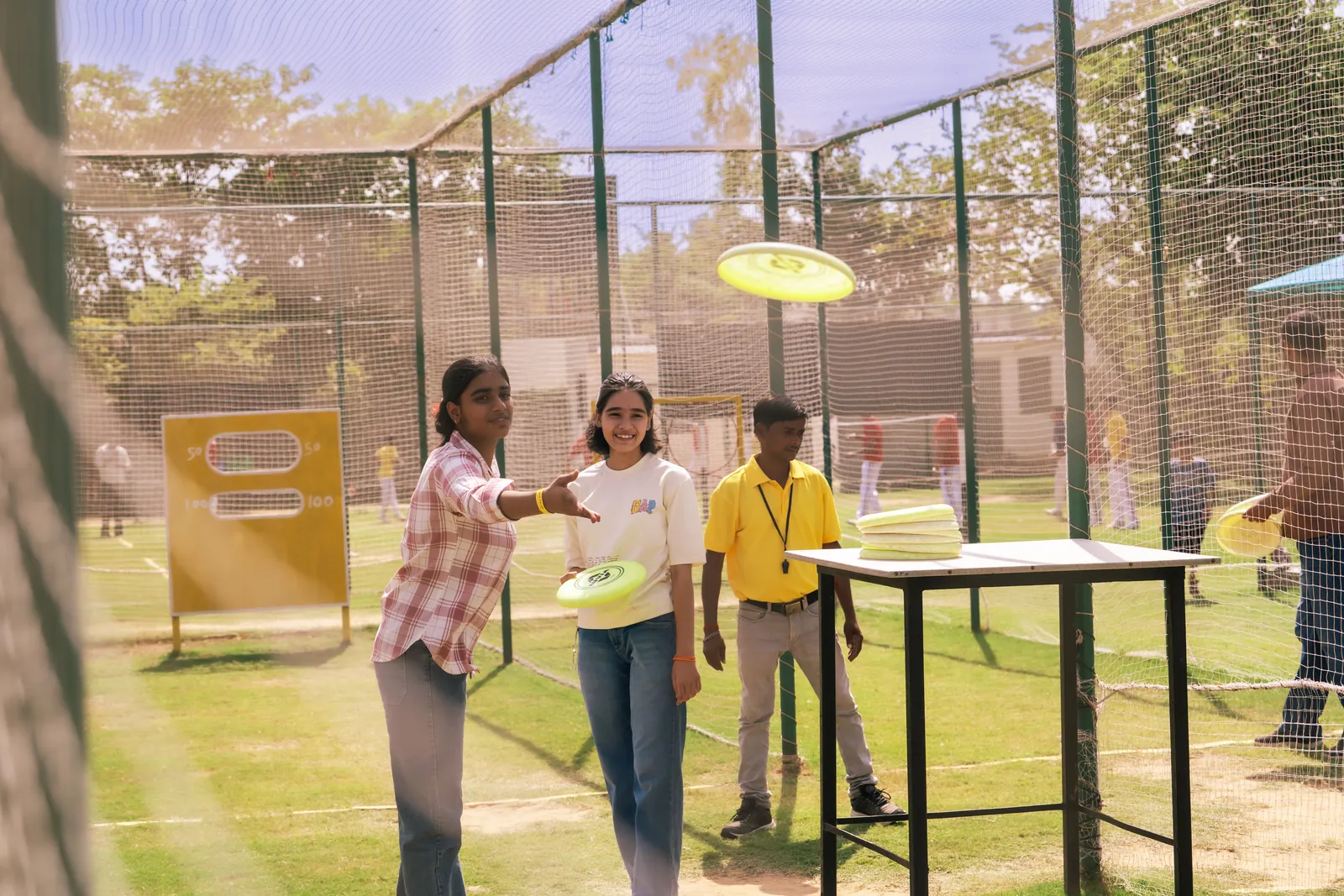 A woman throwing a yellow frisbee disc inside the netted sports arena at Pratapgarh Farms, with two guests and a staff member waiting beyond her