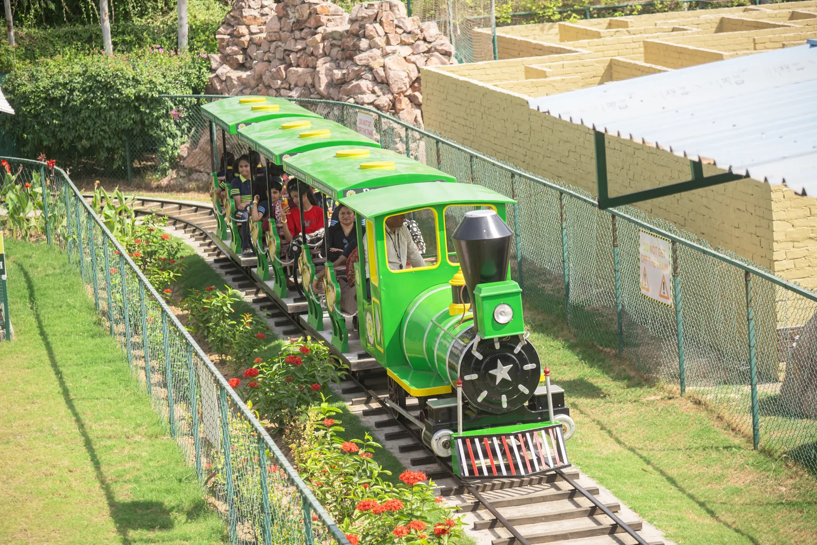 The green Toy Train Ride running along its track beside a lawn and a stone wall at Pratapgarh Farms