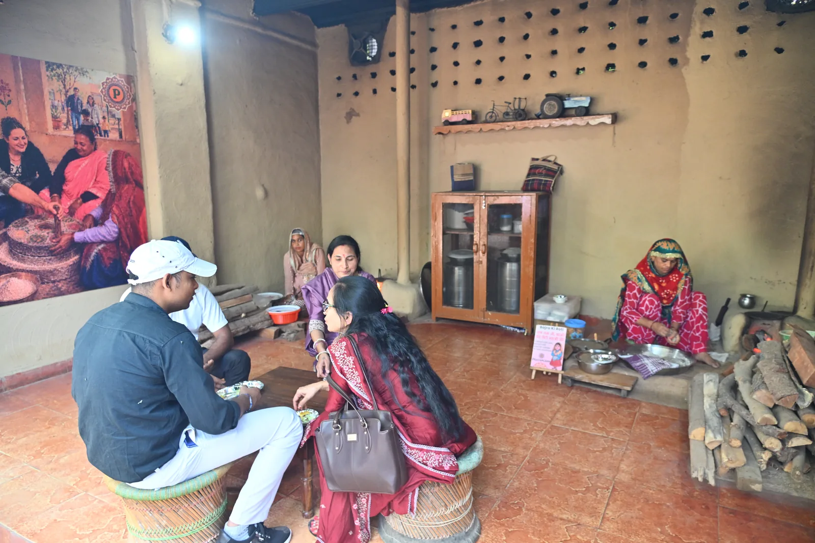 Guests seated on woven stools inside a mud-walled village hut, with a woman cooking at the chulha and a wooden grain chest against the wall