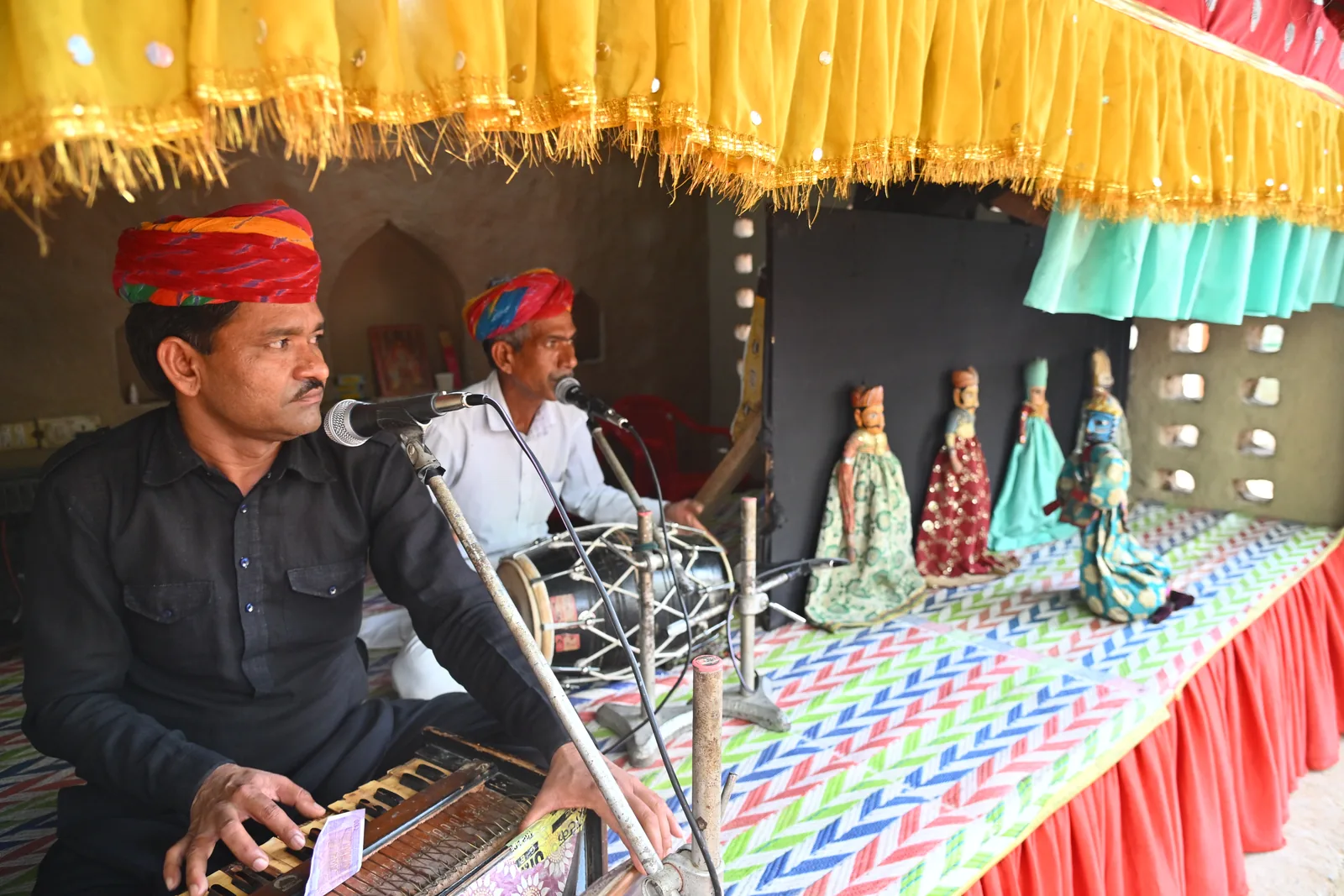 Two folk musicians in red turbans playing the harmonium and the dholak beside a row of colourful kathputli puppets