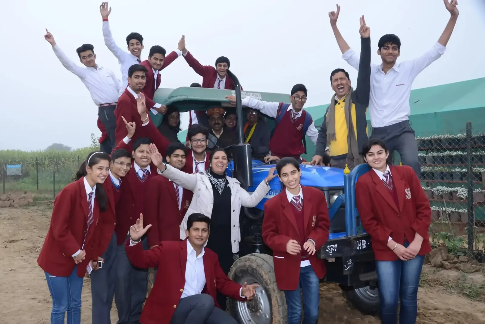 School students in maroon blazers riding a tractor trolley with their arms raised in the air at Pratapgarh Farms