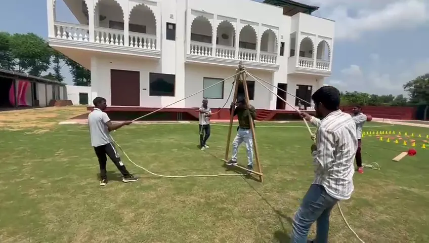 Colleagues pulling ropes from four sides to raise a wooden tripod frame on the lawn