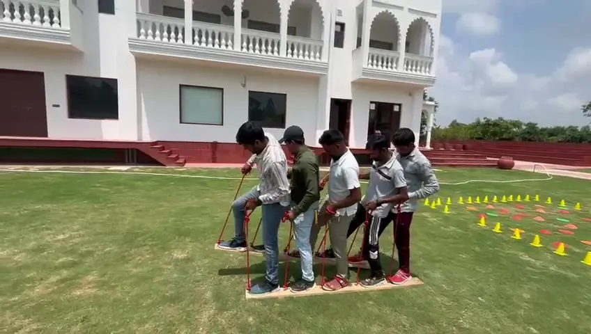 Five colleagues balancing on linked wooden planks, stepping together during a team walk drill on the lawn