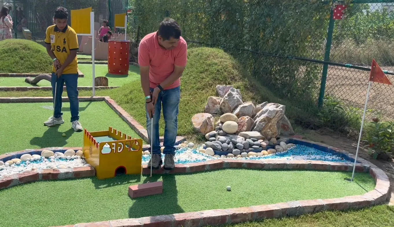 Two boys putting on the mini golf course, with a flag and a stone-lined water channel beside the green