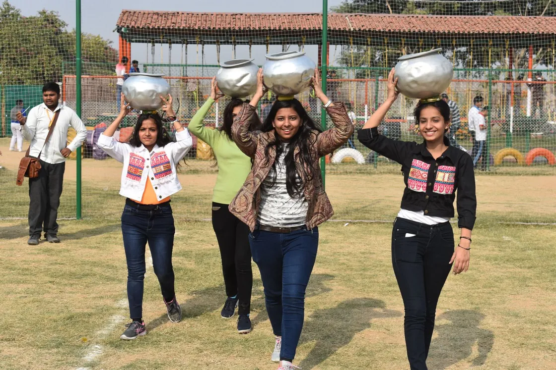 Four women walking across the field with shining metal water pots balanced on their heads
