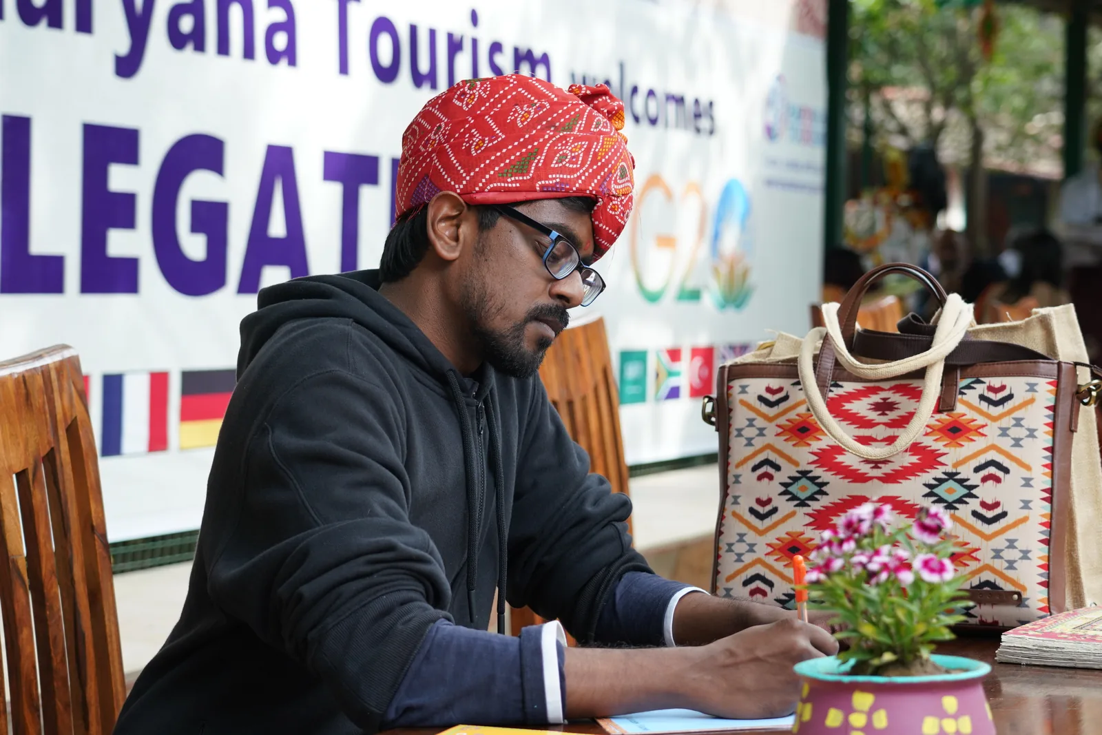A delegate in a red bandhani pagdi signing the visitor register at a desk under a Haryana Tourism welcome banner
