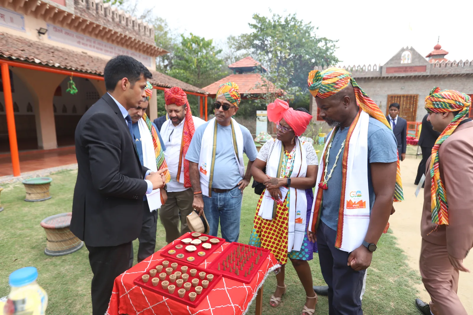 A host presenting trays of round ghee and jaggery products to delegates in colourful pagdis at the Vedic Gaugram