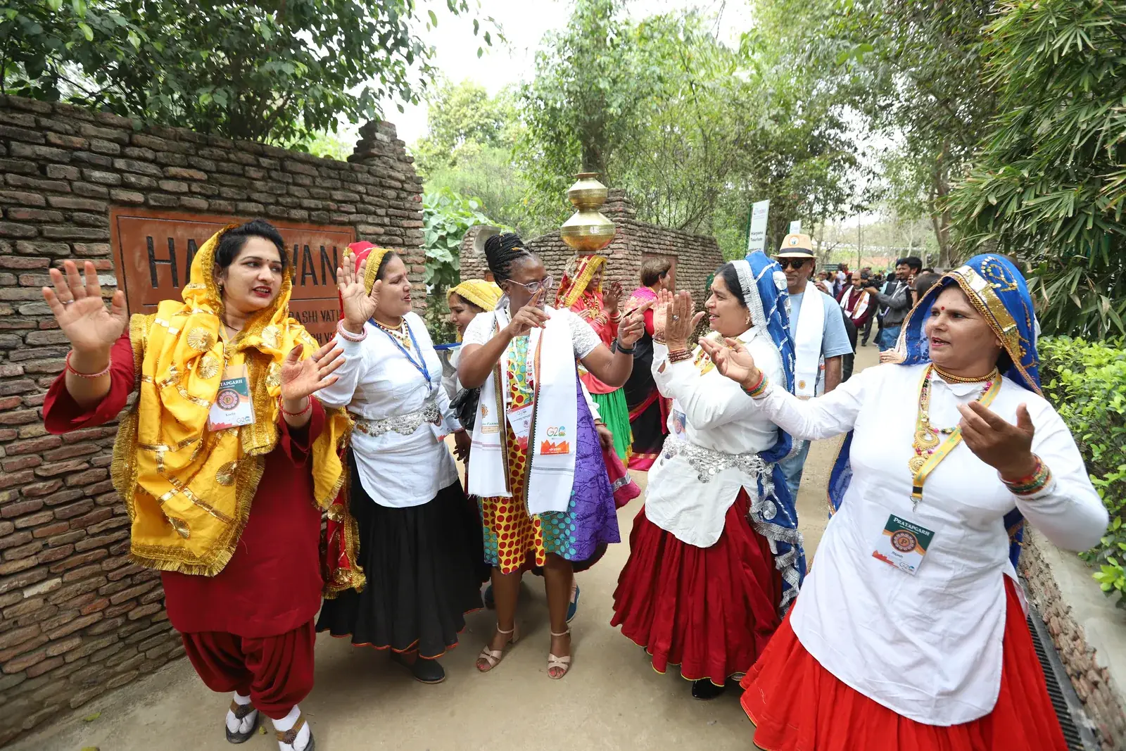 Women in bright Haryanvi dress with a brass kalash, singing and clapping to welcome the G20 delegation inside the farm