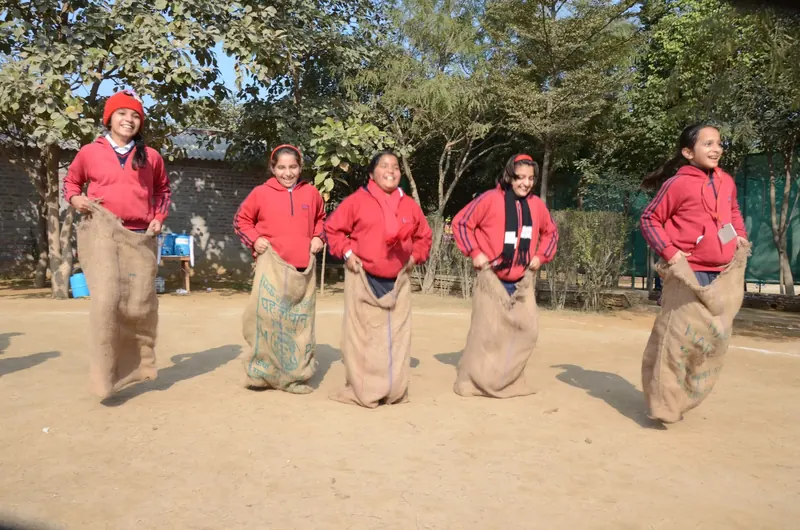 Students in red hoodies hopping in a sack race across a dirt ground