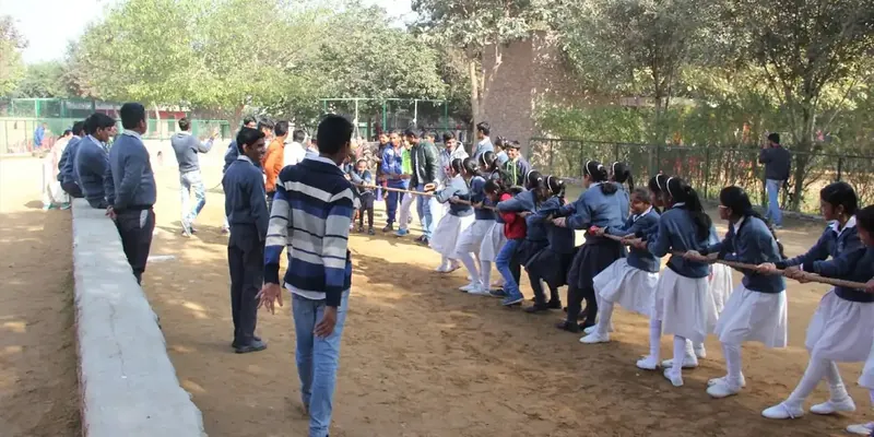 A long line of students in sweaters and white salwar pulling a rope in a tug-of-war on bare earth
